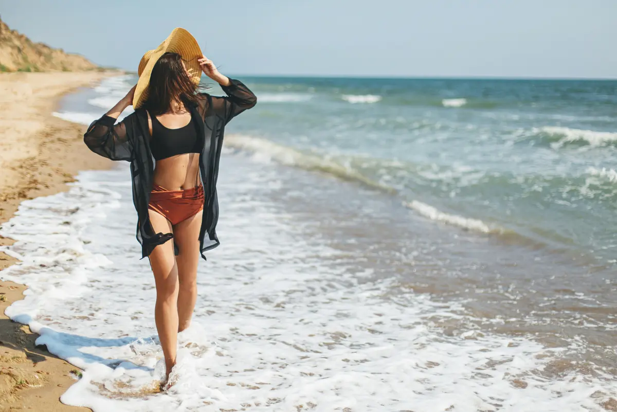 Junge Frau geht am Strand spazieren, sie trägt einen Strohhut, ein schwarzes Hemd und einen Bikini
