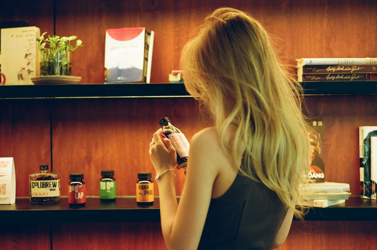 Rückansicht einer Frau, die vor einem Regal steht und eine kleine Parfumflasche in der Hand hält
