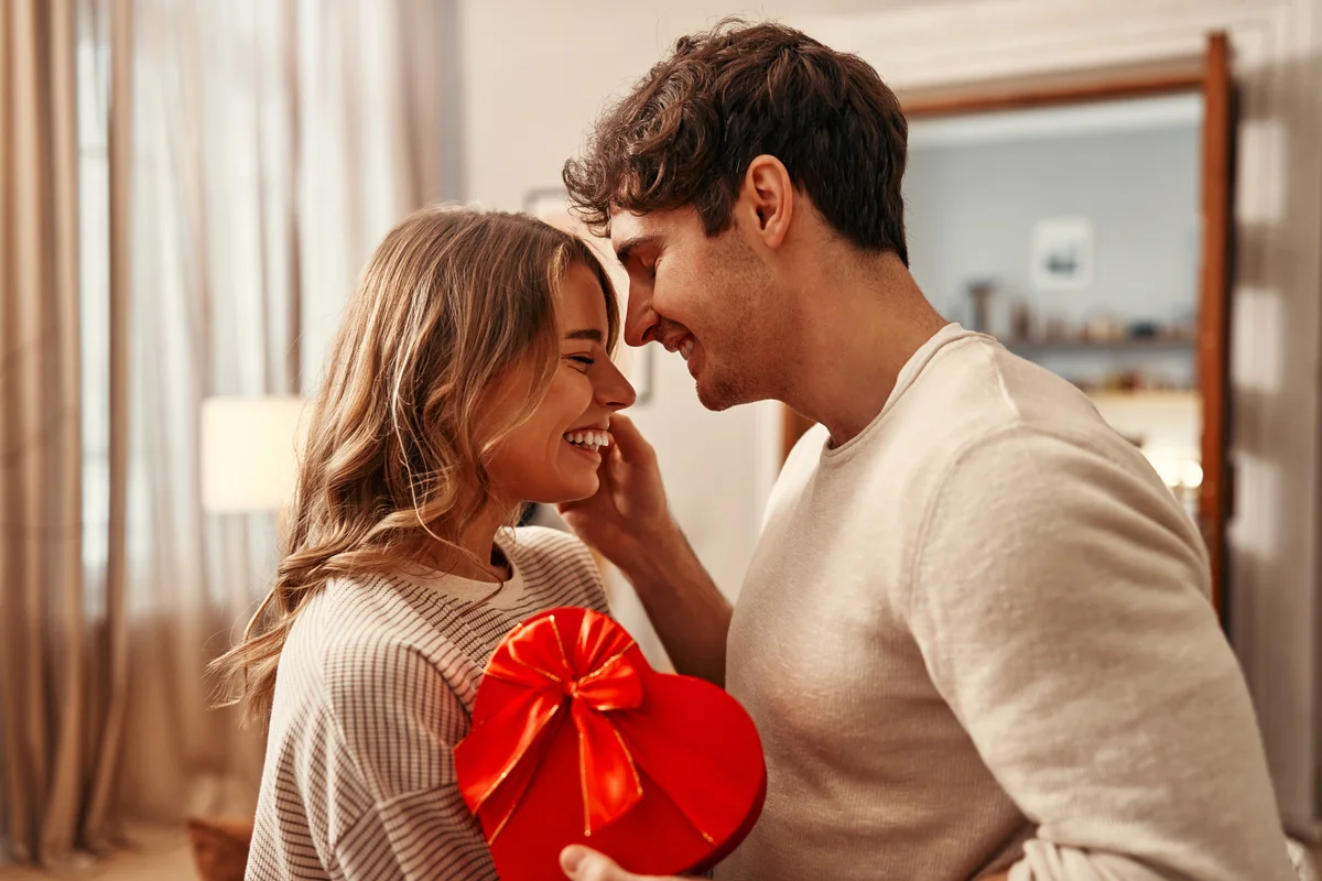 Valentinstag. Verliebtes Paar zu Hause. Er hat eine rote Herschachtel in der Hand.