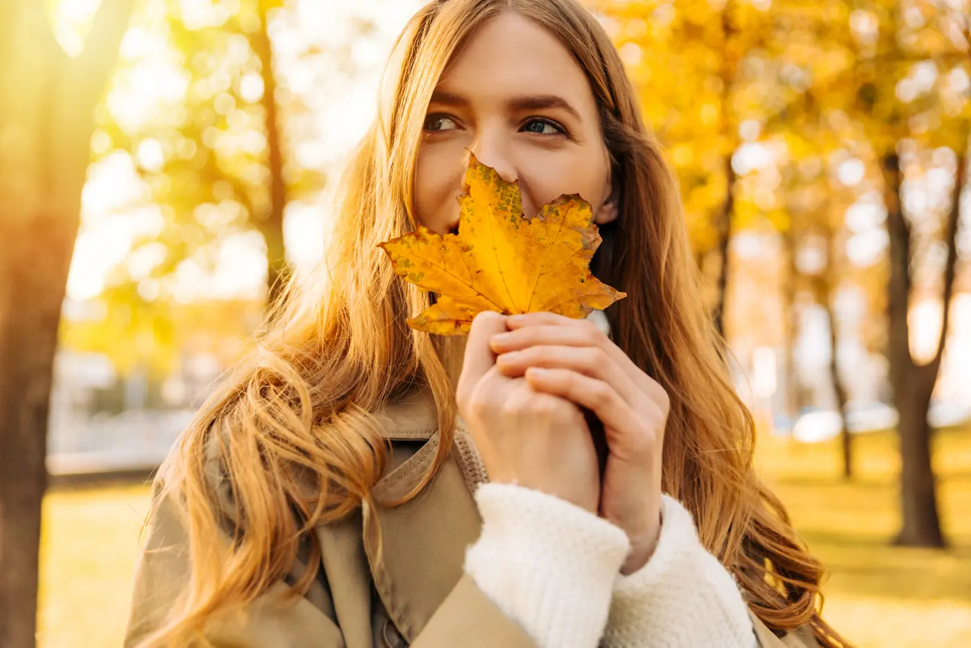 Schöne Frau in einem beigen Regenmantel zeigt ein gelbes Blatt, das von einem Baum gefallen ist.