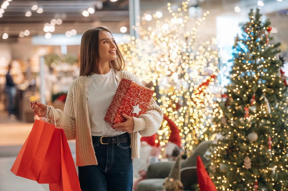 Junge Frau mit roten Einkaufstüten und einem verpackten Geschenk vor einem beleuchteten Weihnachtsbaum