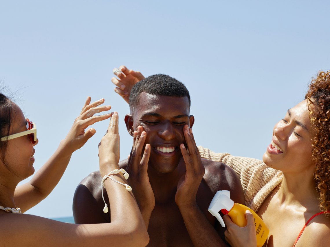 Zwei Frauen tragen einem lachenden Mann am Strand Sonnencreme fürs Gesicht auf