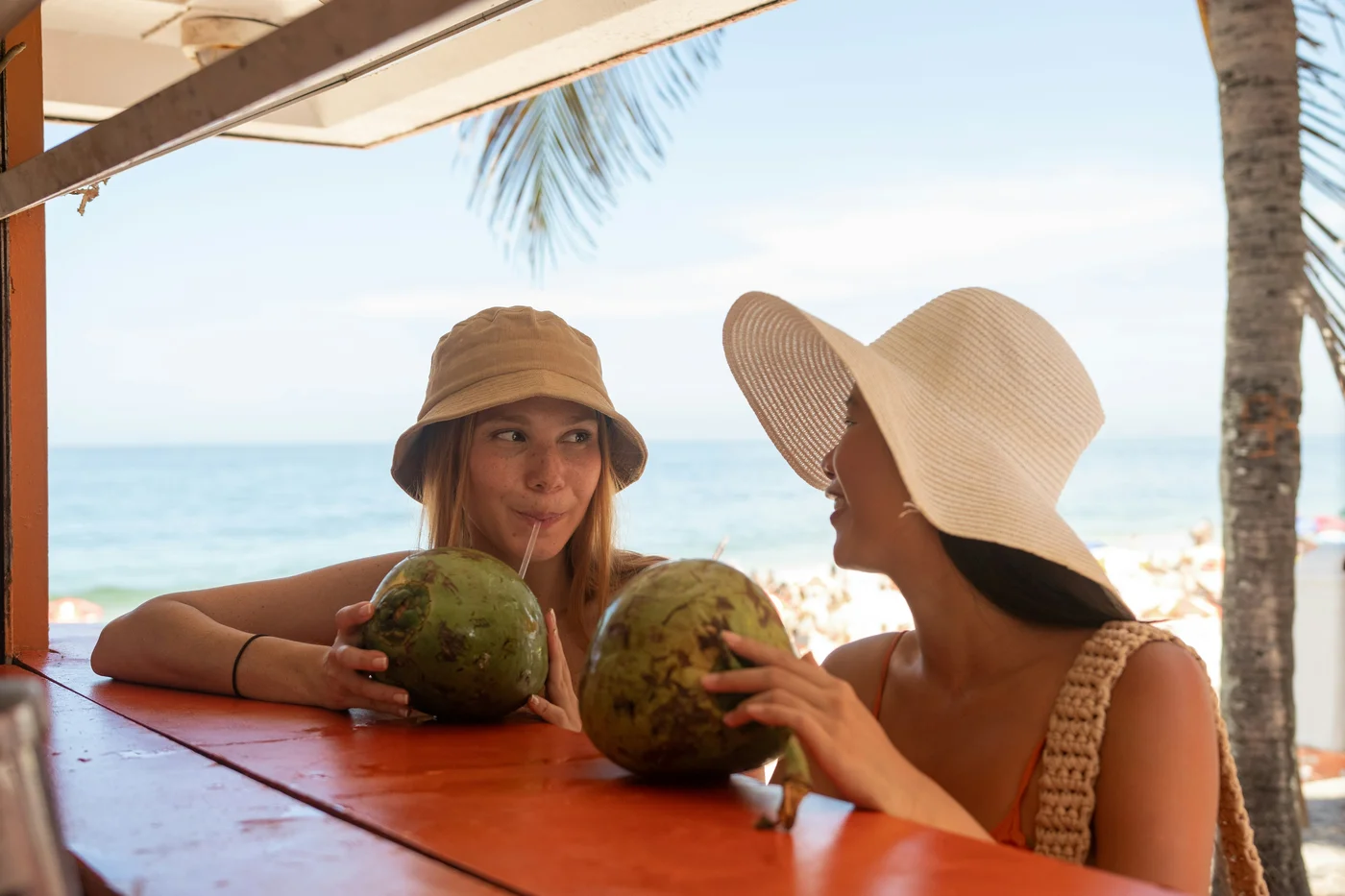 Zwei Frauen mit Strohhüten sitzen unter einer Palapa an einem Strand und trinken aus grünen Kokosnüssen