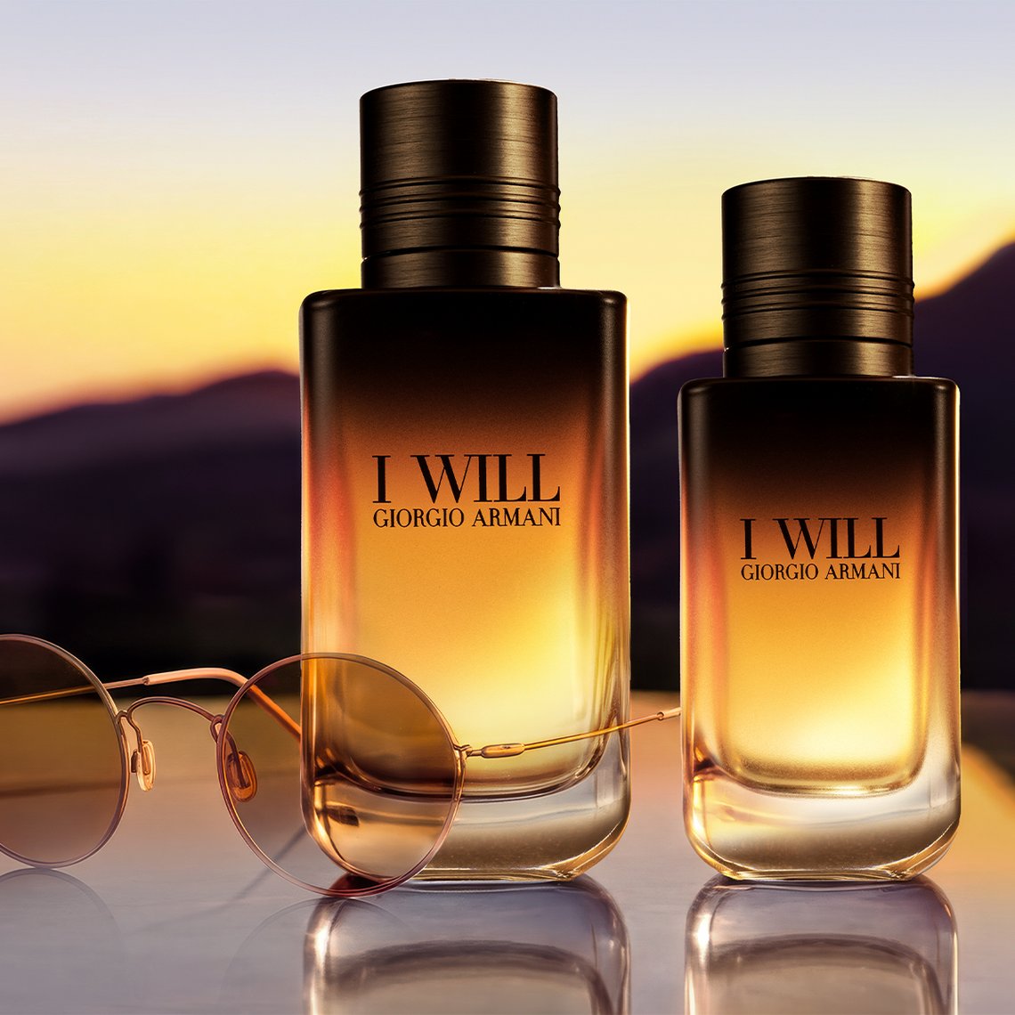Zwei Giorgio Armani I Will Parfümflakons in unterschiedlichen Größen neben einer Sonnenbrille auf spiegelnder Fläche vor Bergsilhouette im Sonnenuntergang