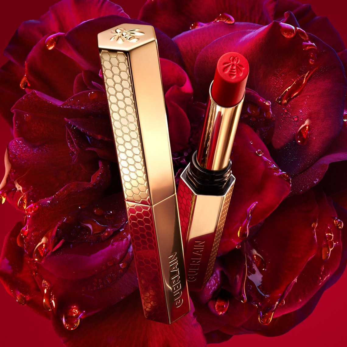 Geöffneter Guerlain KissKiss Lippenstift mit Bienen-Prägung und goldener Hülse, arrangiert vor einer großen, dunkelroten Rose mit Wassertropfen.