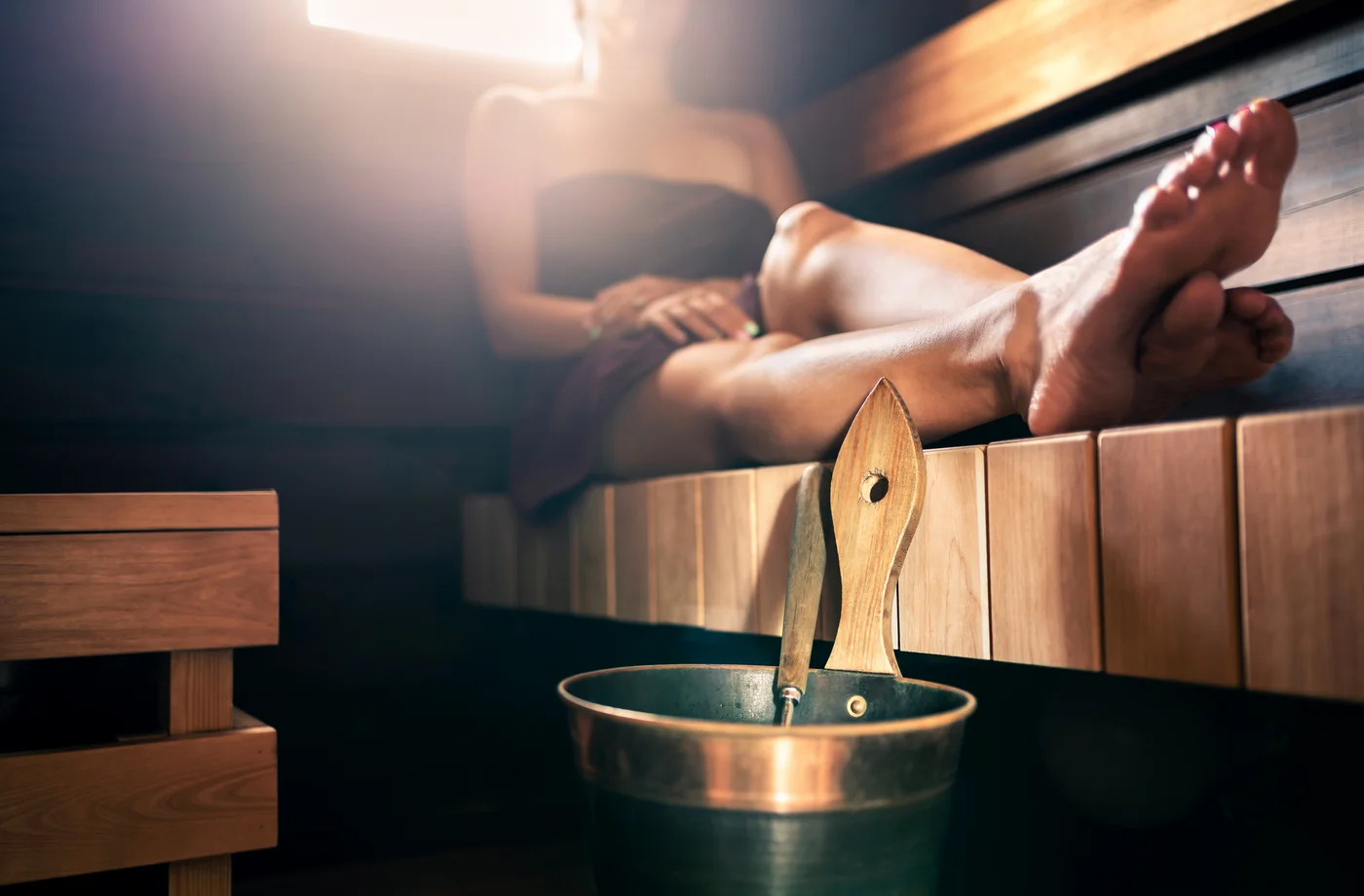 Frau liegt entspannt auf der Holzbank einer Sauna, im Vordergrund ein Saunakübel mit Aufgusskelle