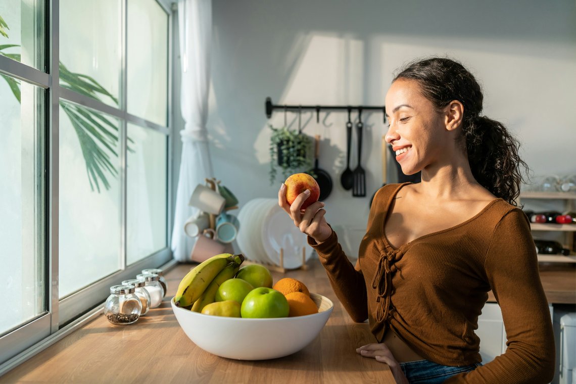 Lächelnde Frau steht in einer hellen Küche vor einer Schale mit frischem Obst und hält einen Apfel in der Hand.