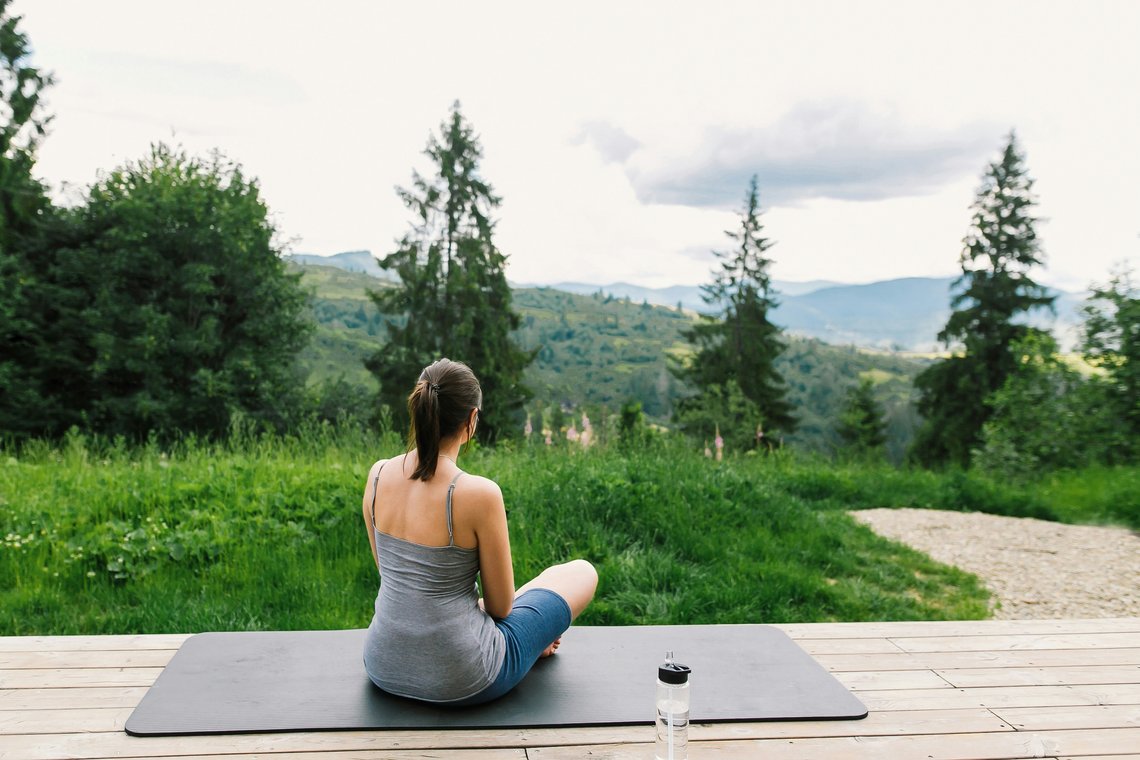 Frau sitzt entspannt auf einer Yogamatte auf einer Holzterrasse und blickt auf eine grüne Berglandschaft.