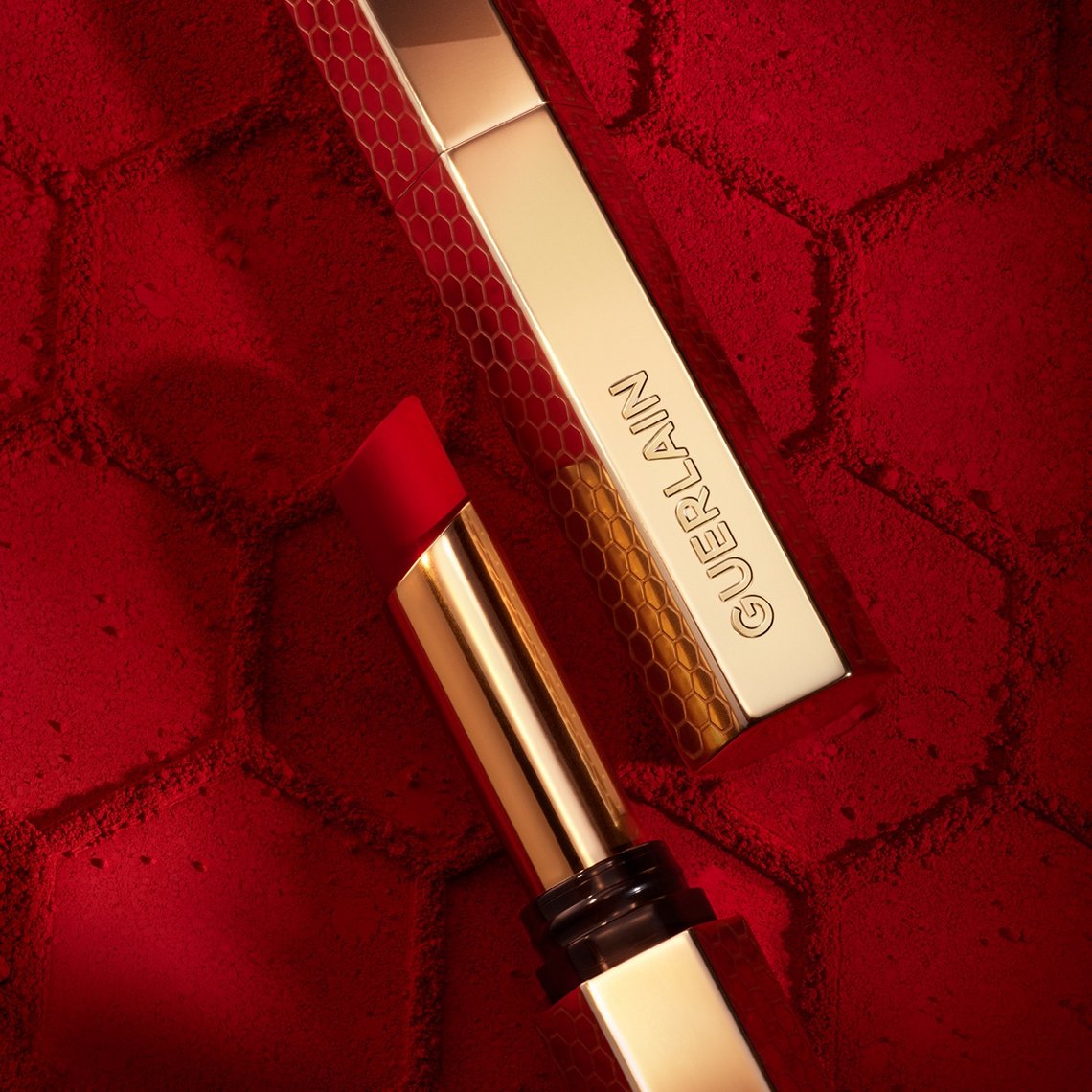 Guerlain KissKiss Lippenstift in hellem Rot mit geöffneter, goldener Hülse auf strukturiertem, rotem Hintergrund mit Wabenmuster.