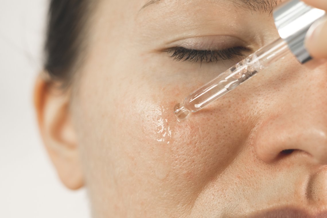 Nahaufnahme eines Gesichts mit geschlossenem Auge, aus einer Pipette fällt ein klarer Serumtropfen auf die Wange unterhalb des Auges
