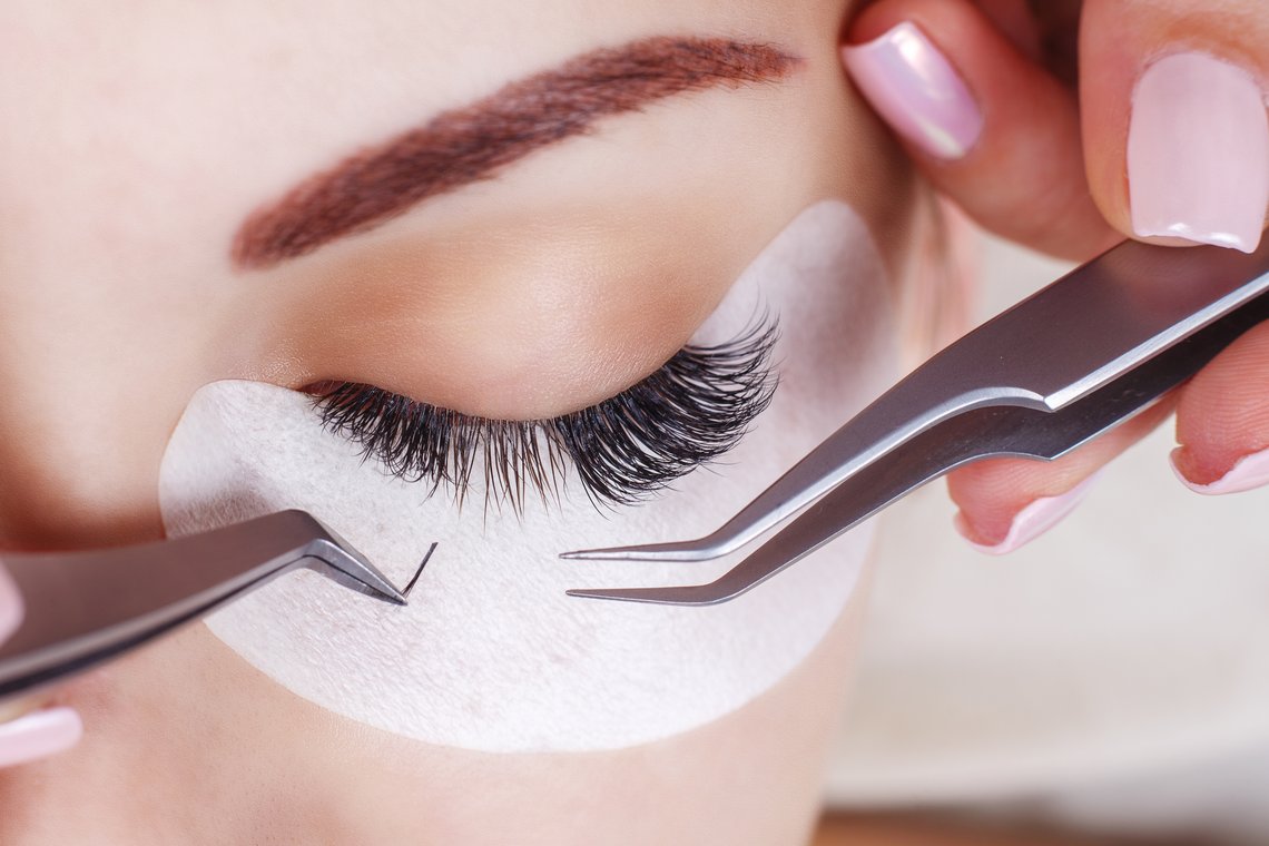 Nahaufnahme eines geschlossenen Auges mit Augenpad, an dem zwei Pinzetten einzelne Wimpern ansetzen