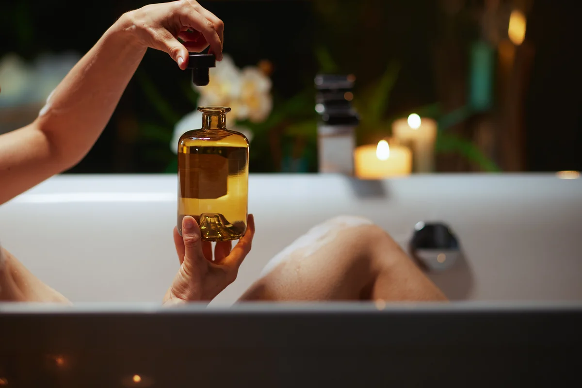 Frau in der Badewanne hält eine Flasche Badeöl bei Kerzenlicht