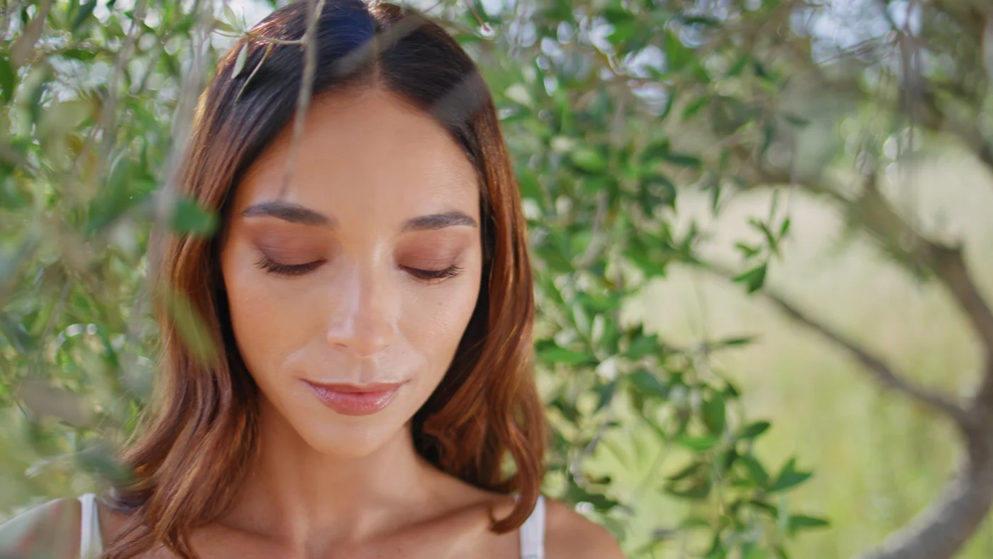 Nahaufnahme auf das Gesicht einer jungen Frau mit geschlossenen Augen, im Hintergrund Blätter eines Baumes