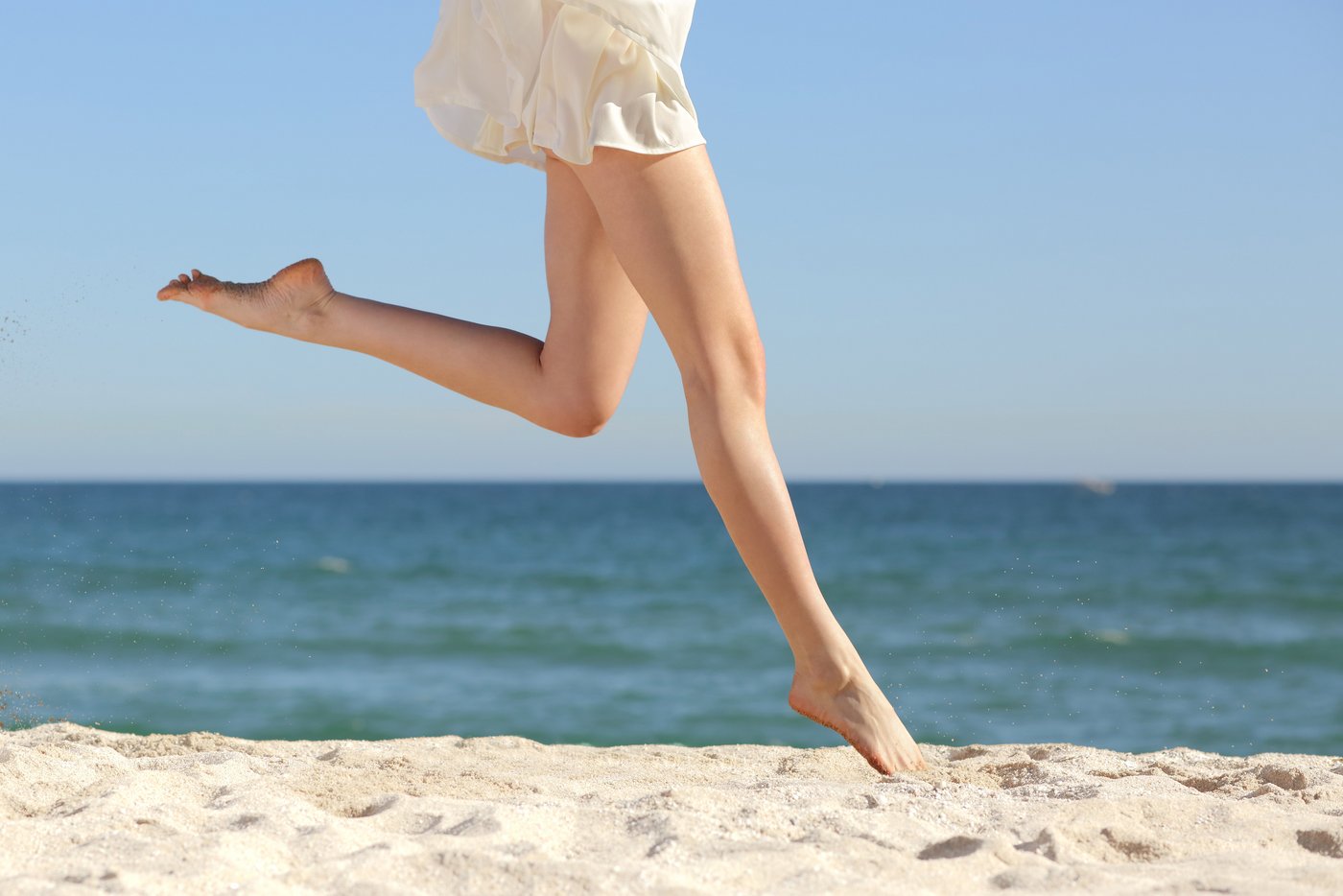 Frau springt am Strand durch den Sand mit nackten Beinen
