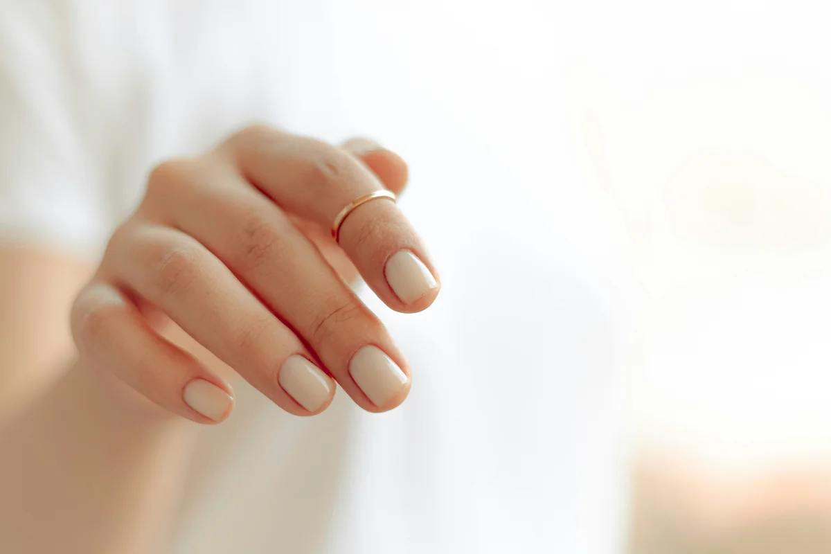 Kurze Gelnägel in einem cremigen Nude-Ton an einer Hand mit schmalem Goldring