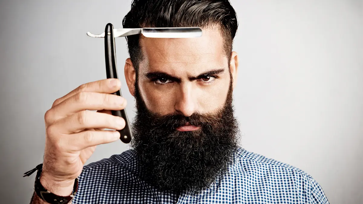 Ein Mann mit Vollbart hält ein Rasiermesser vor sein Gesicht