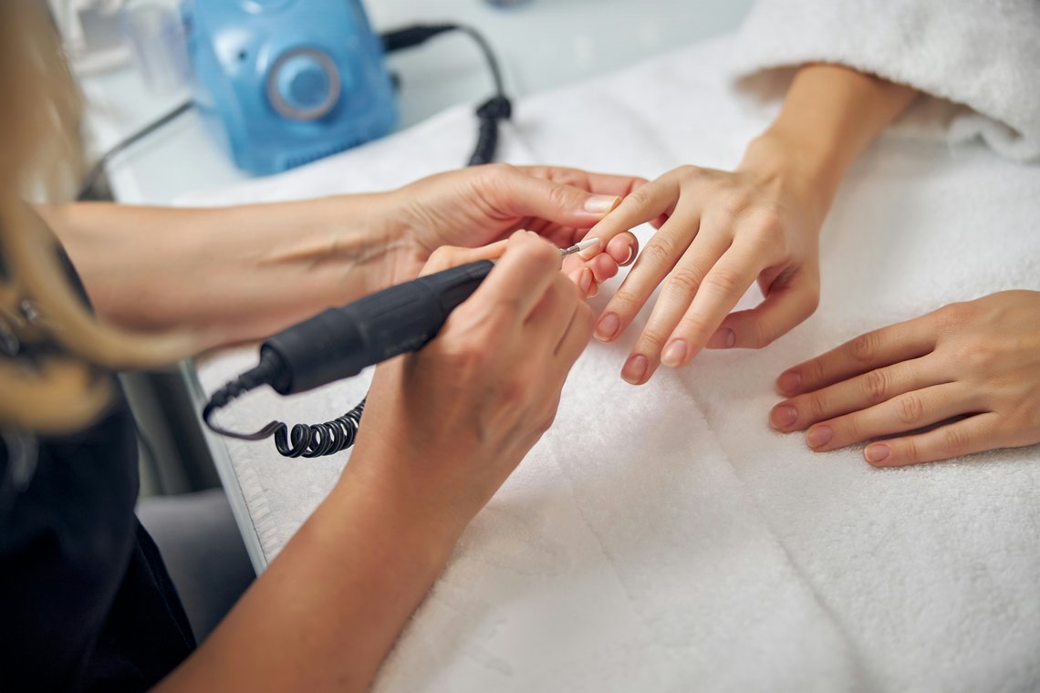 Nageldesignerin im Nagelstudio bearbeitet die Nagelhaut einer Kundin mit einem elektrischen Nagelfräser.