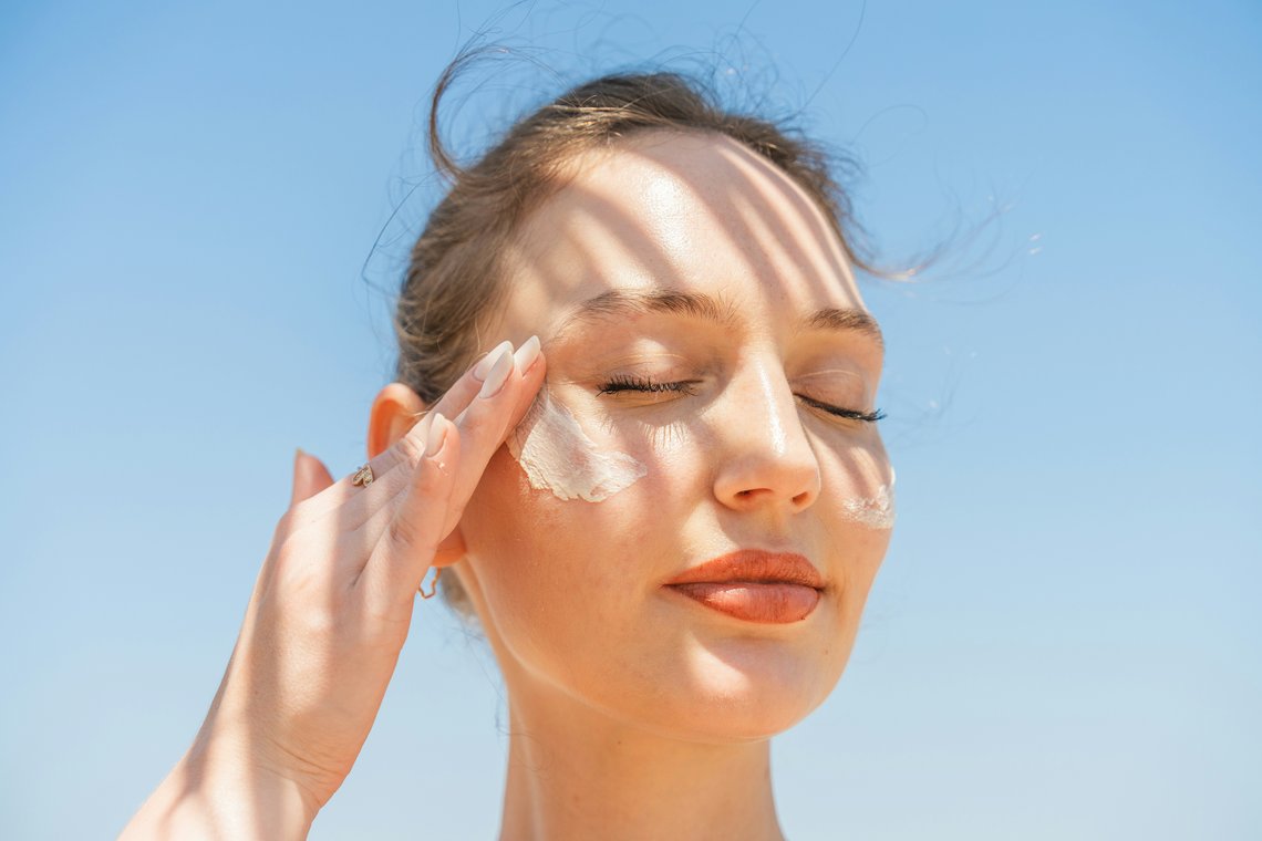 Frau mit geschlossenen Augen trägt vor blauem Himmel Sonnencreme auf die Wange auf.
