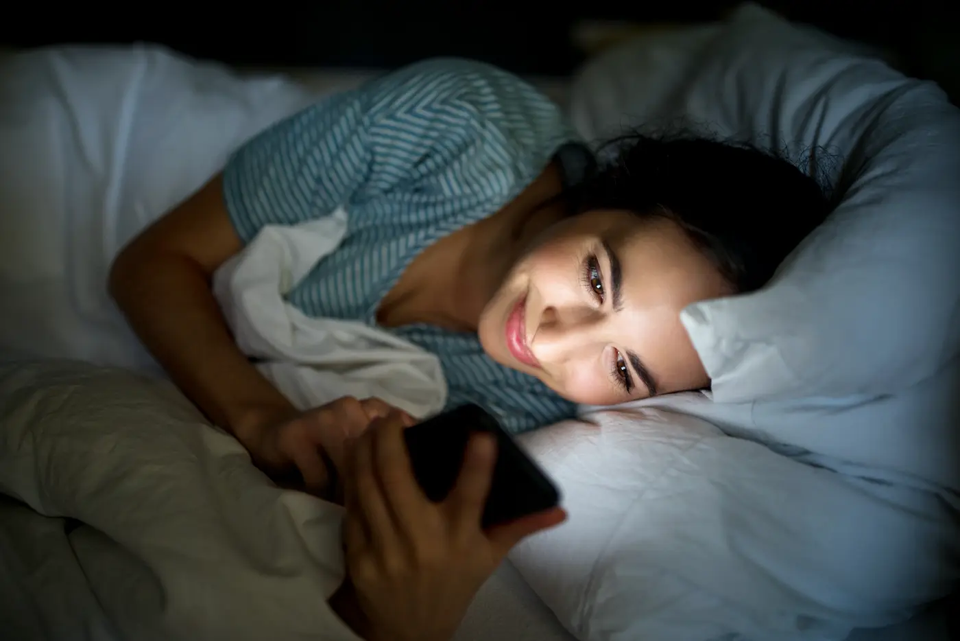 Frau liegt im Bett und sieht auf ihr Handy, beleuchtet vom blauen Licht des Displays