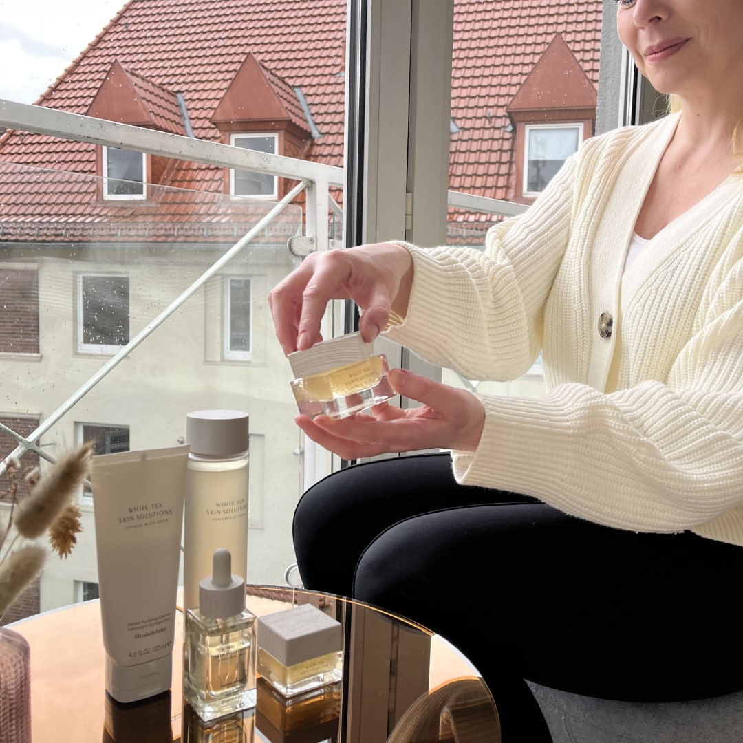 Die Produkte der White Tea Skin Solutions stehen auf einem kleinen Tisch vor einem Fenster, die Redakteurin hält ein Produkt in der Hand