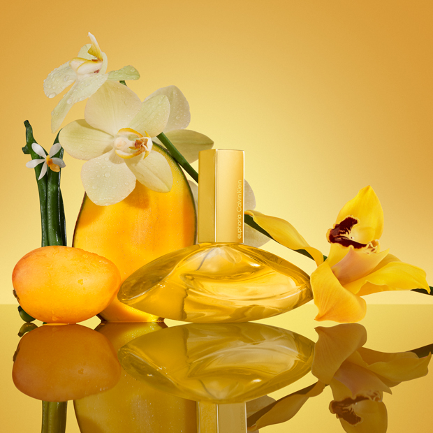 Calvin Klein Euphoria Elixir Duft in goldenem Flakon, inszeniert mit Früchten, Blüten und Vanille auf spiegelnder Oberfläche