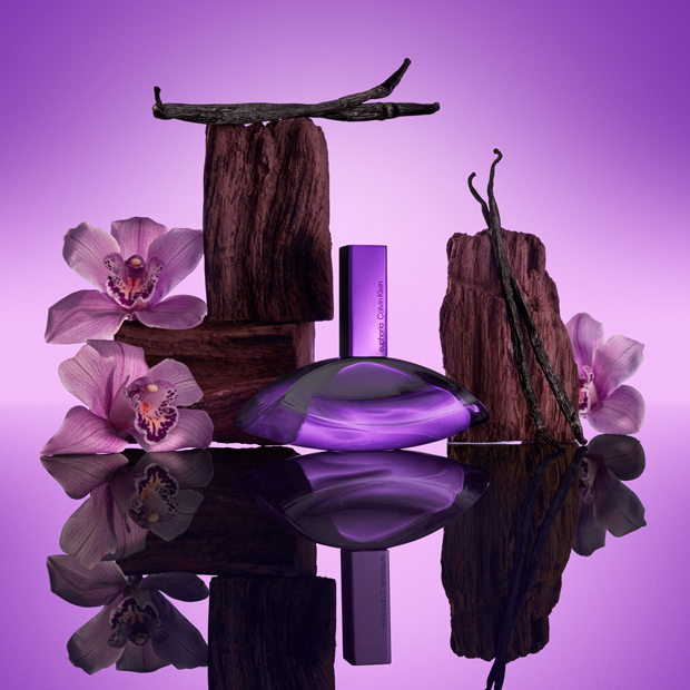 Calvin Klein Euphoria Elixir Duft in violettem Flakon, inszeniert mit Vanilleschoten, Holz und Orchideen auf spiegelnder Oberfläche