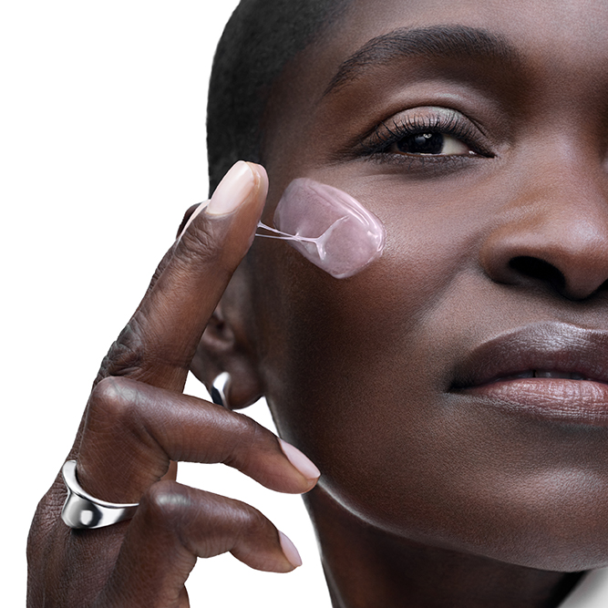 Nahaufnahme einer Frau mit dunkler Haut, die eine rosafarbene, gelartige Pflegecreme sanft auf ihre Wange aufträgt